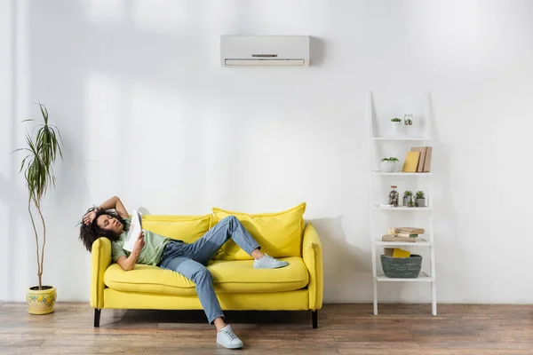 african american woman waving with newspaper while lying on yellow couch and suffering from heat